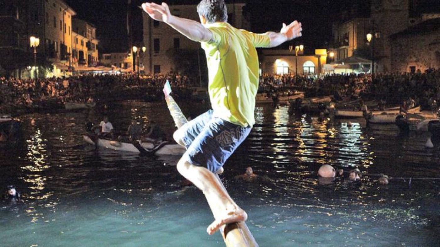 Un tuffatore avanza in equilibrio sul palo della cuccagna, di notte, sul porto di Lazise