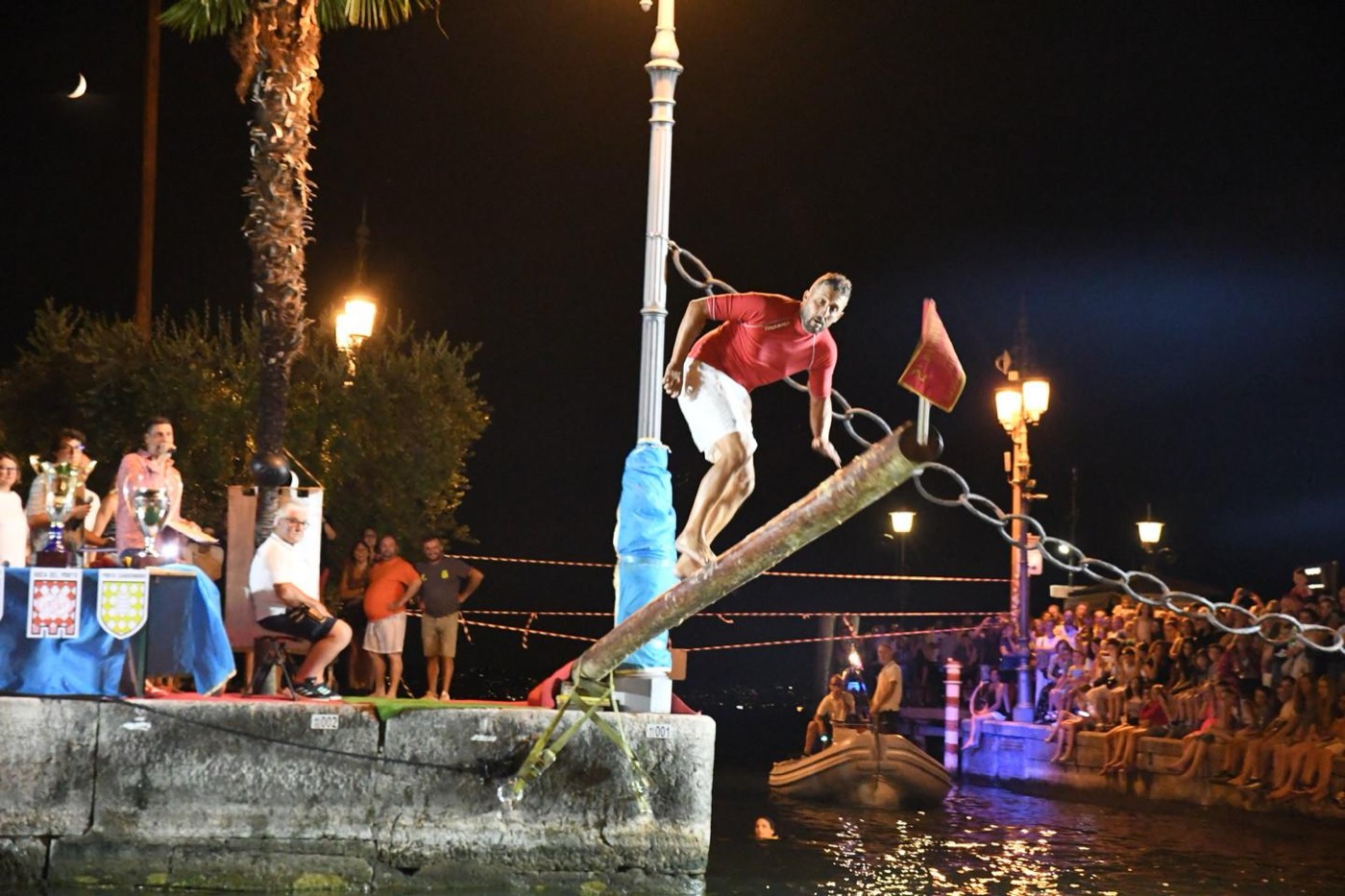 Un concorrente in equilibrio sul palo della cuccagna raggiunge la bandiera di notte, sul Porto Vecchio di Lazise