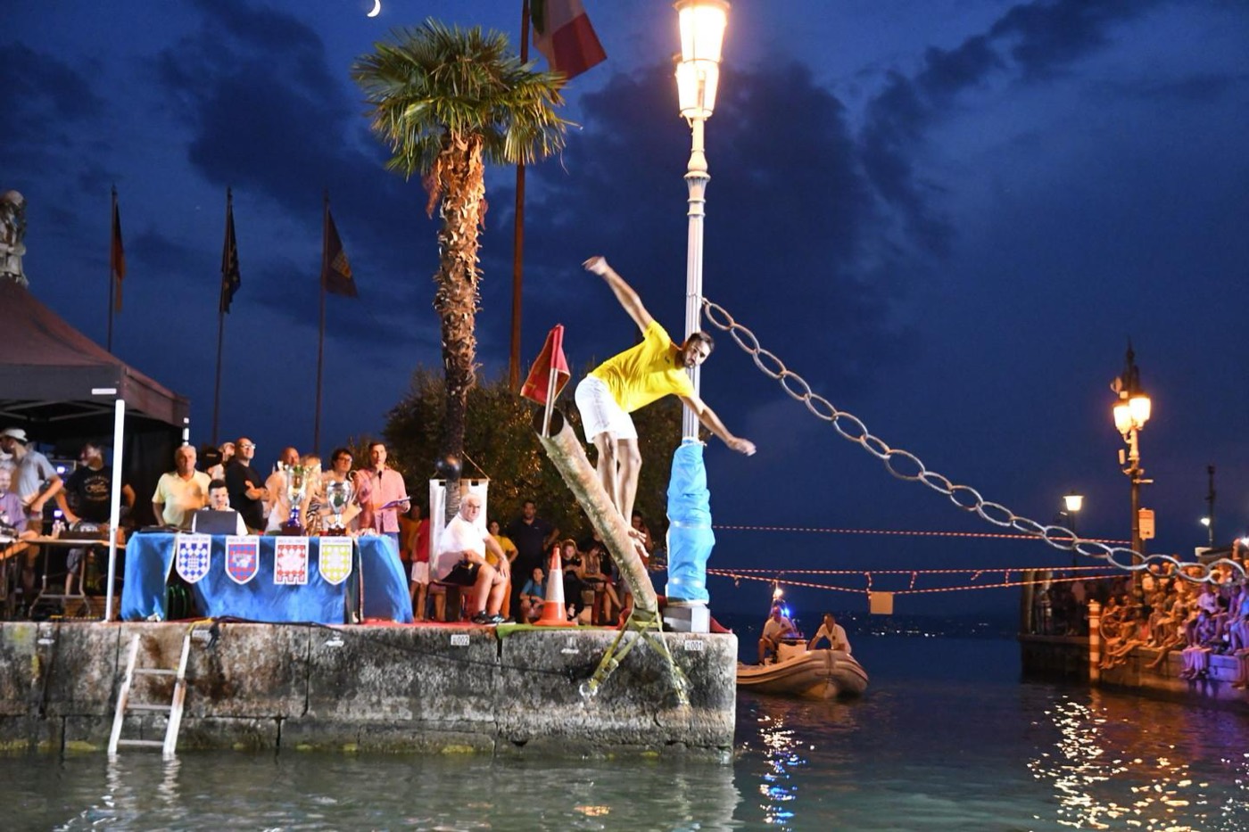 Un tuffatore in equilibrio sul palo della cuccagna al tramonto, tra la folla del porto di Lazise