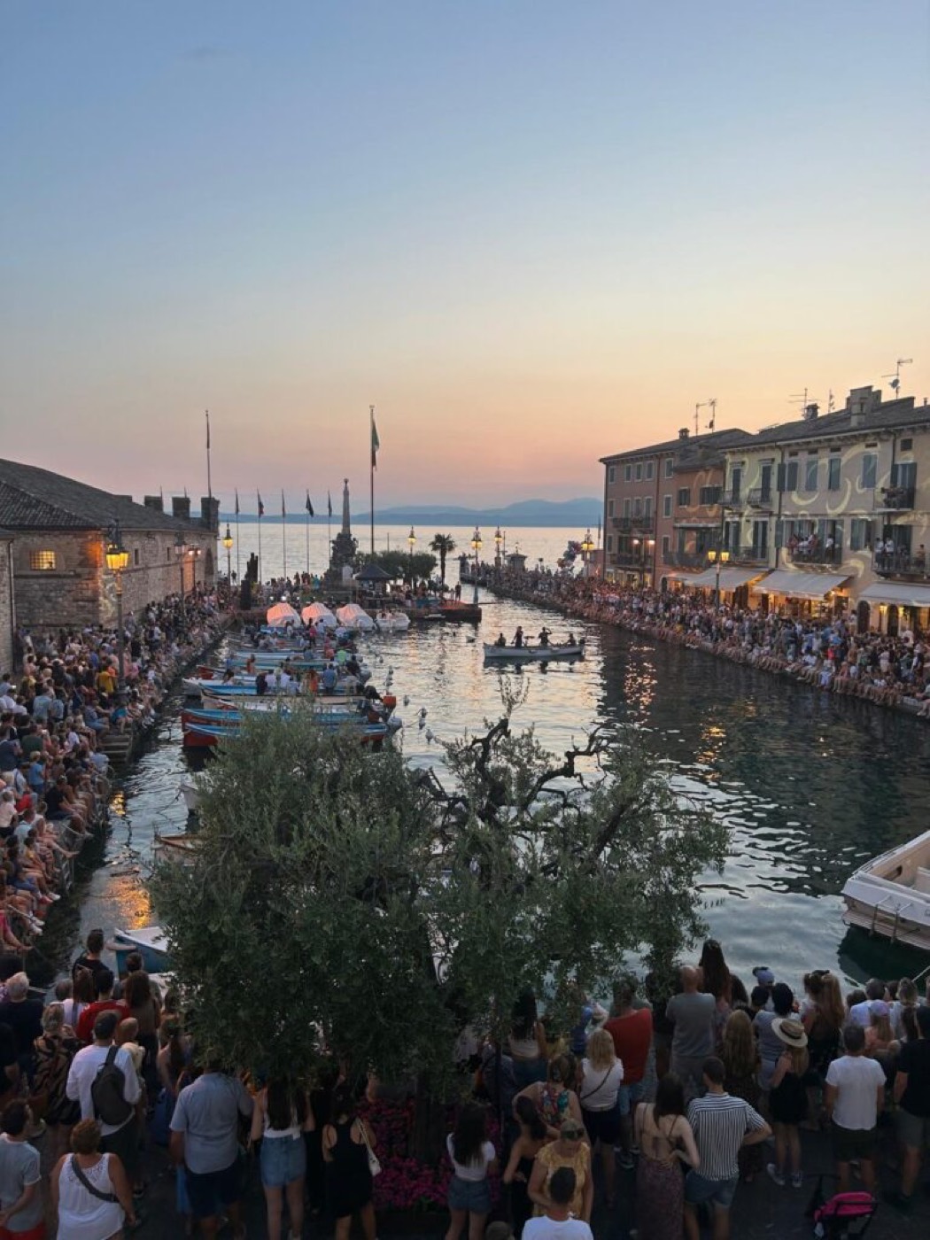 Il Porto Vecchio di Lazise gremito di pubblico al tramonto, con il palo teso sull'acqua